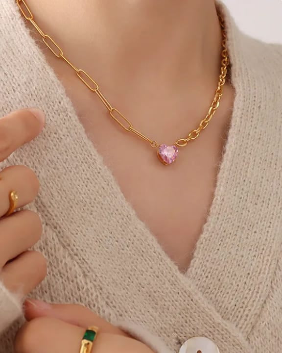 Gold necklace with a pink heart pendant worn by a person in a beige sweater.