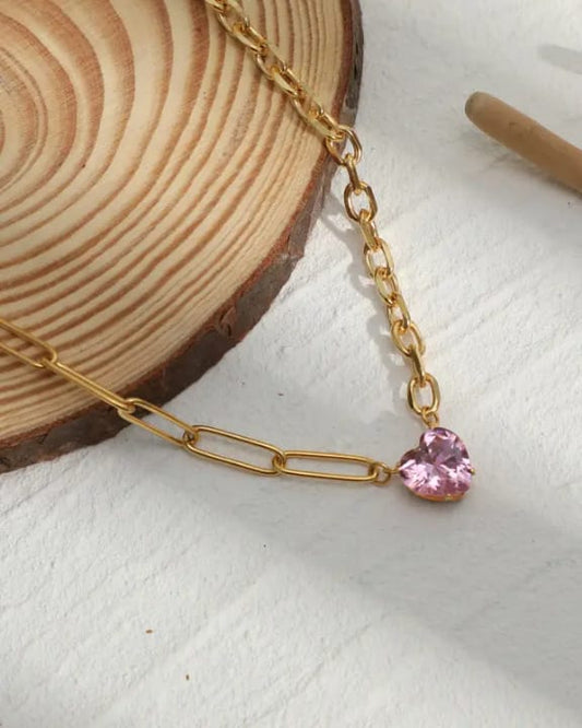 Gold chain necklace with a pink heart-shaped pendant on a wooden and white surface.