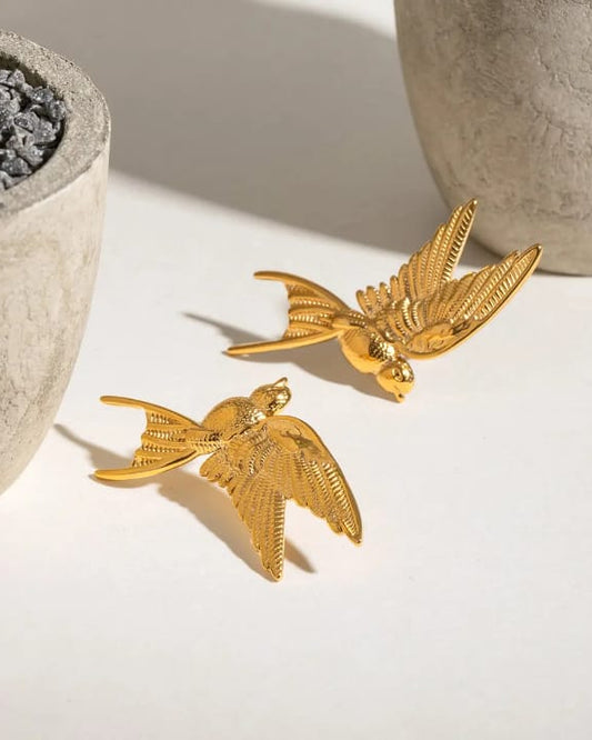 Gold bird-shaped earrings on a light surface with decorative pots in the background