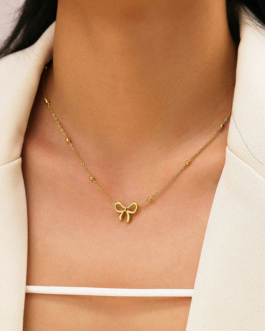 Gold necklace with a bow pendant worn by a person in a white outfit.